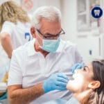 A patient receiving dental treatment from a dentist in a clinic, with a dental assistant in the background.